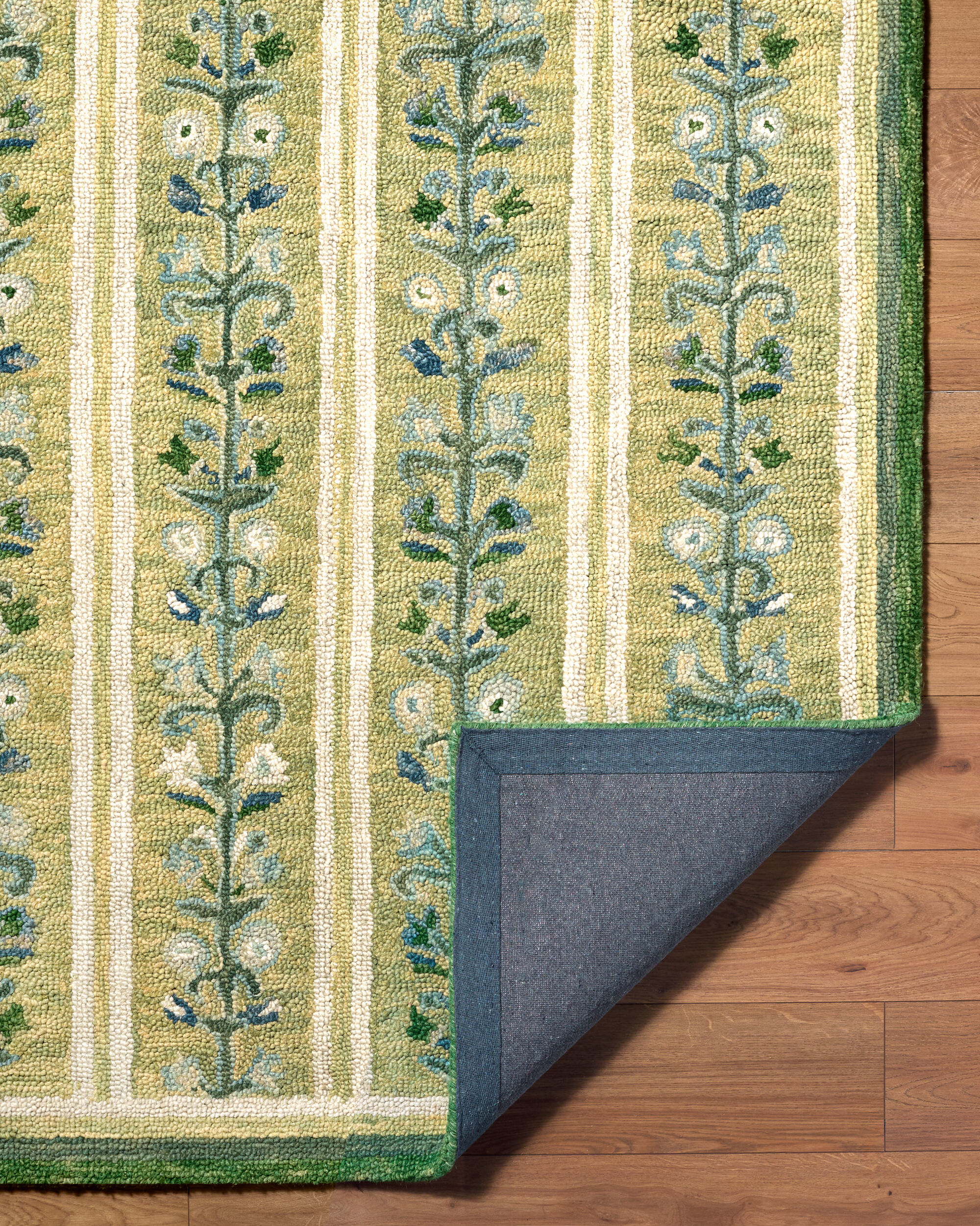 detail of green rug with repeating blue and cream floral vine motifs and ivory stripes with a corner turned up to show the back of the rug