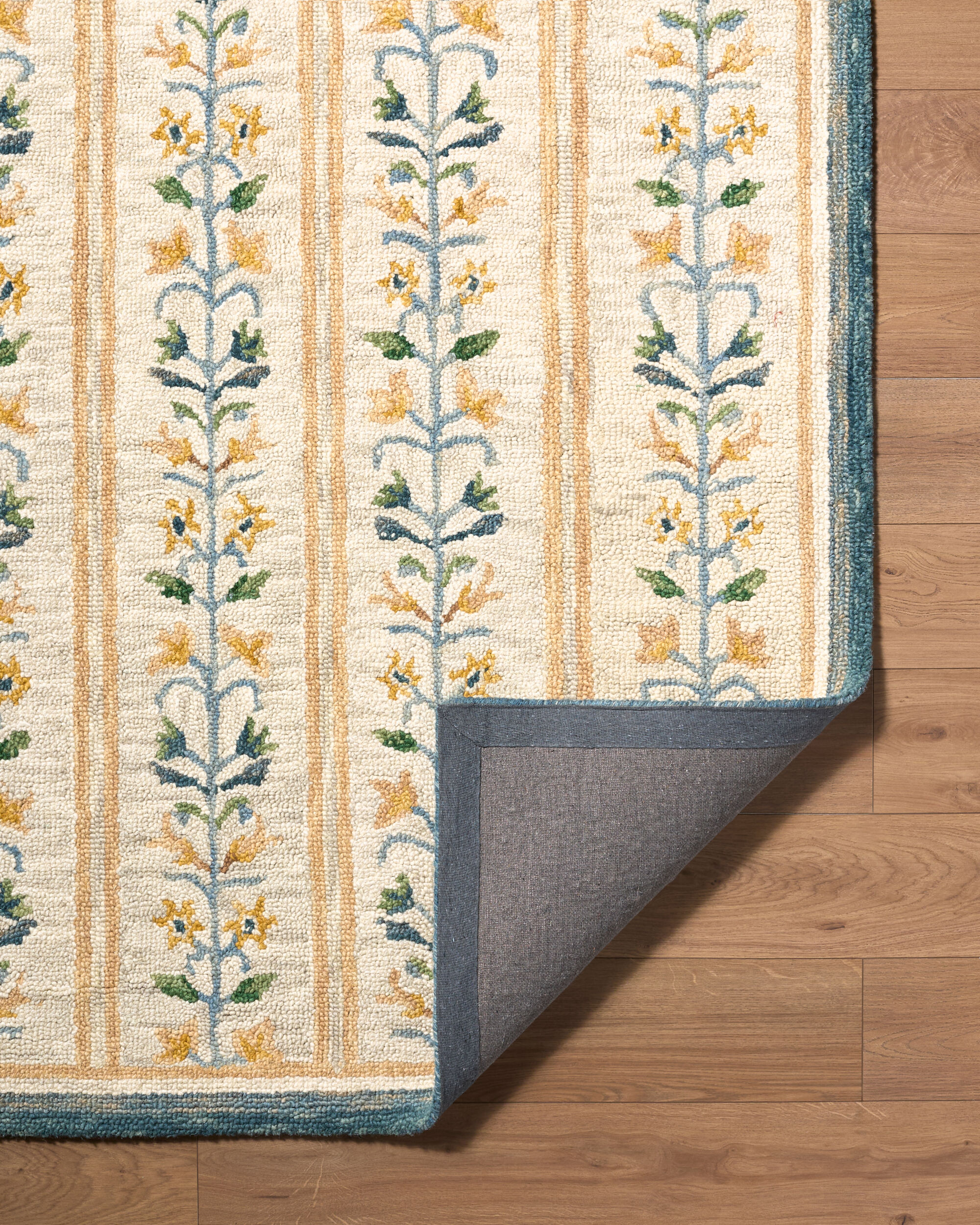 detail of hand tufted ivory rug with repeating blue and gold floral vine motifs, gold stripes, and a dark blue border with a corner turned up to show the back of the rug