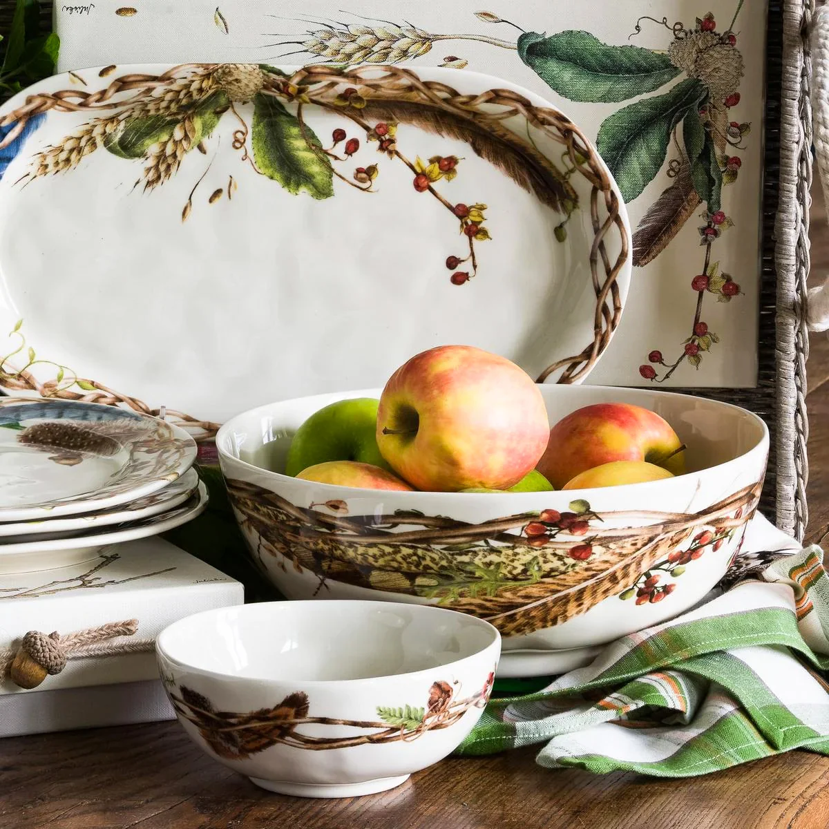 serving pieces with painterly motifs of flora, fauna, and found woodland treasures shown stacked and with a bowl of apples
