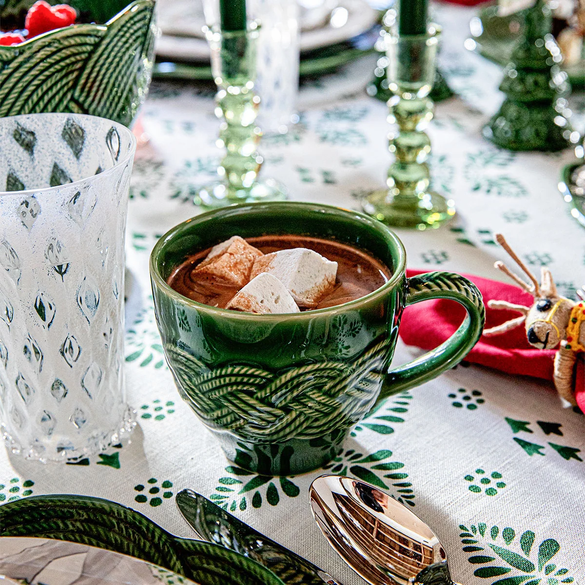dark green mug with intricate, interwoven braiding filled with cocoa and marshmallows on a holiday table