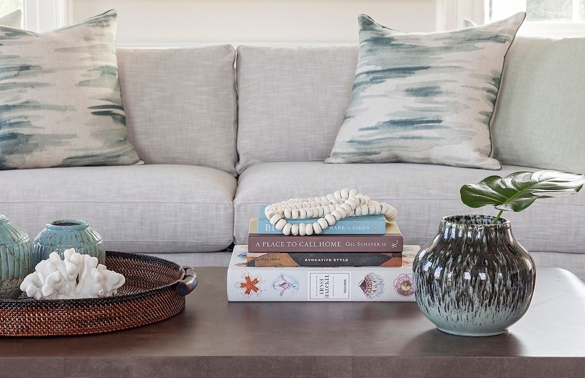 Quiet luxury interior design on display in a Charleston living room, with a beige sofa, teal-accented throw pillows, fine ceramic vases and decorative objects, a walnut coffee table, and a decorative stack of coffee table books