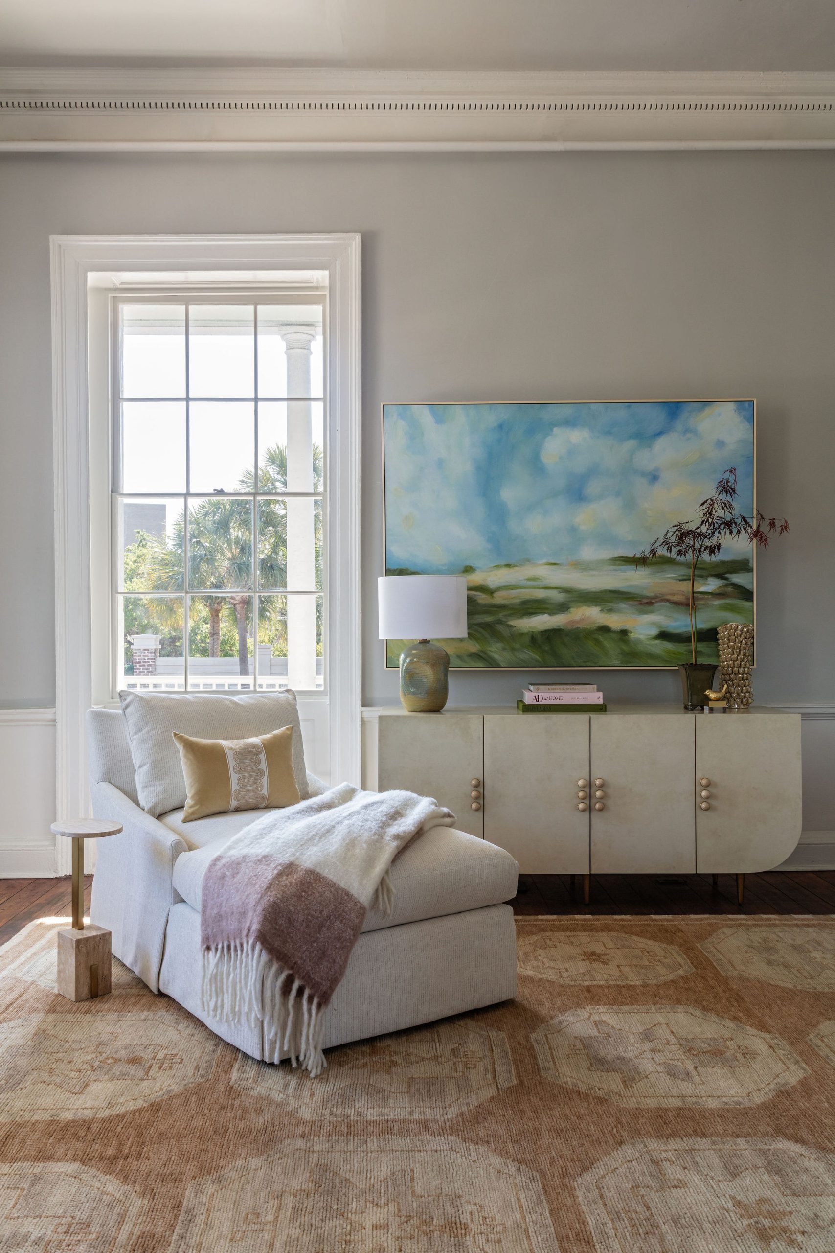lifestyle image of parchment colored buffet with rounded bottom corners and stone hardware, neutral colored chaise with gold pillow, and a blue and green landscape painting