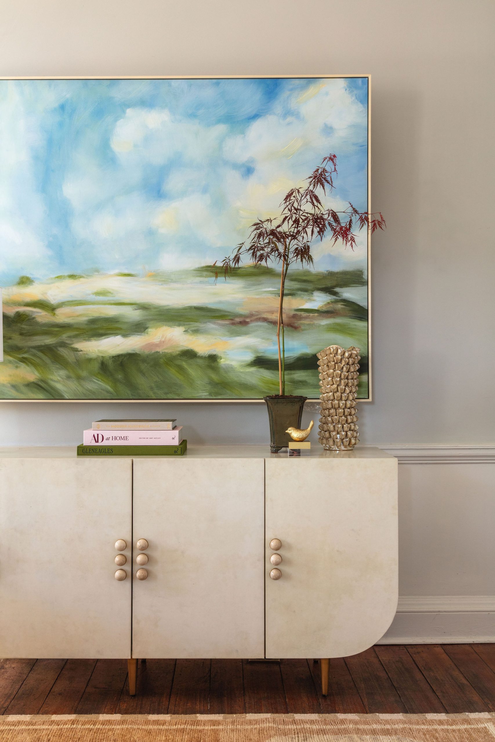 lifestyle image of parchment colored buffet with rounded bottom corners and stone hardware and a blue and green landscape painting