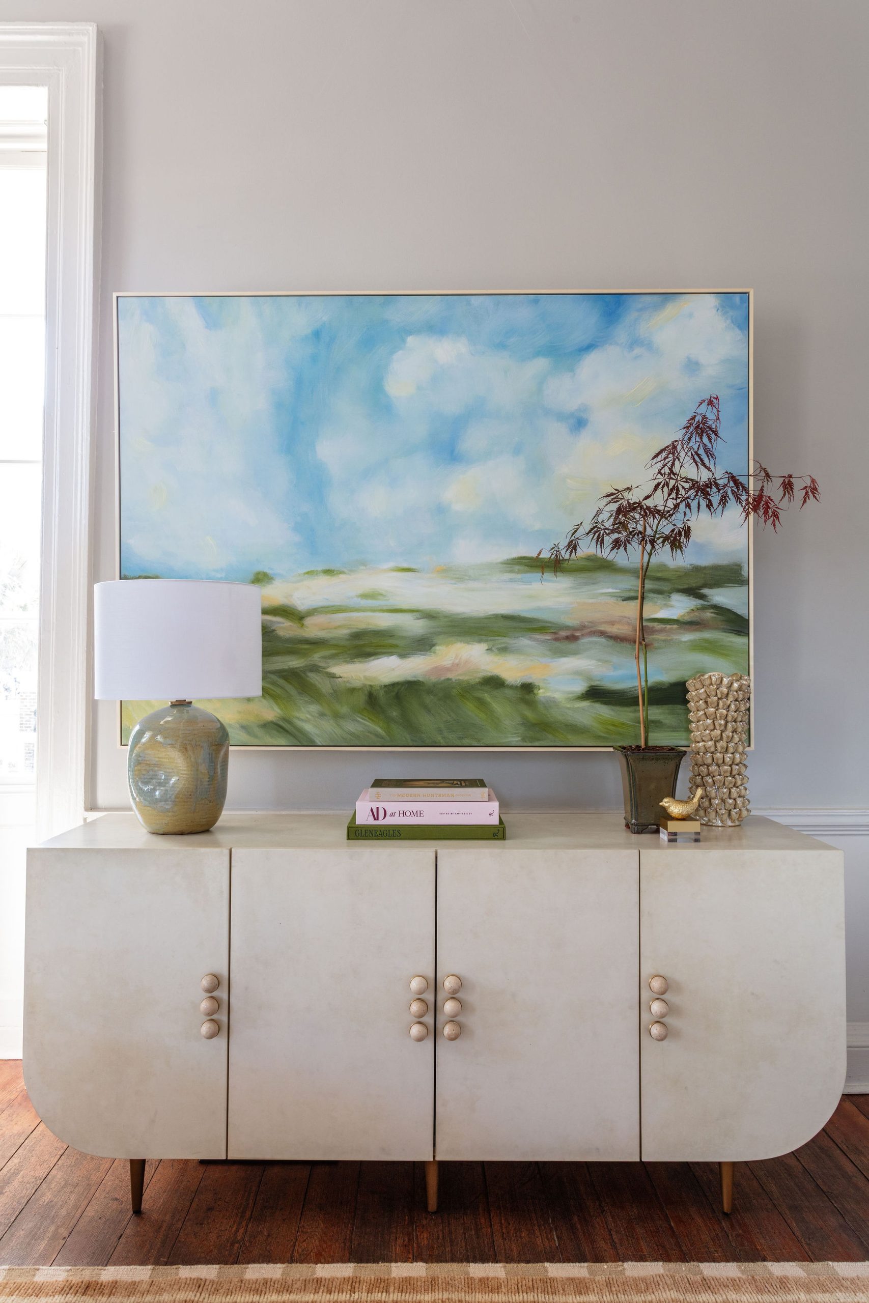 lifestyle image of parchment colored buffet with rounded bottom corners and stone hardware and a blue and green landscape painting