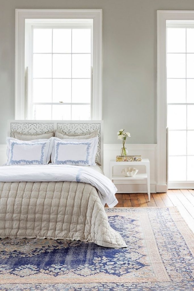 a bedroom designed by the design center of the best furniture store in Charleston, GDC Home, featuring a blue patterned rug, a white bedside table, and bedding including a beige bedspread and white sheets with blue accents