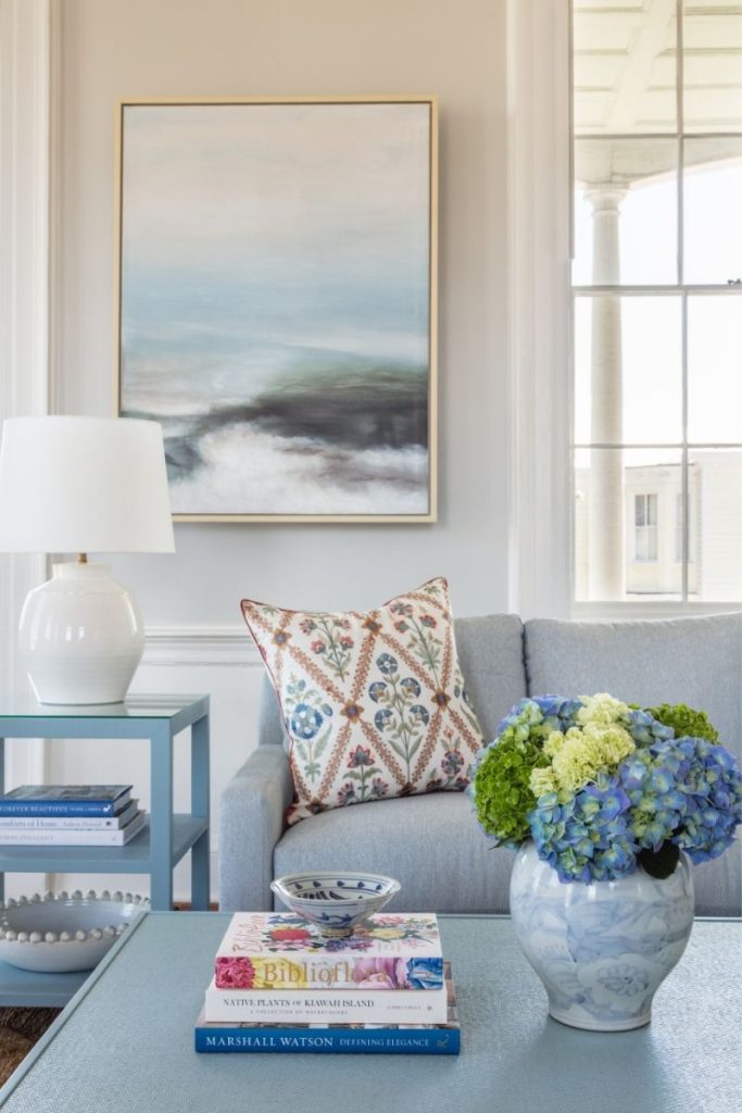 living room with coastal interior design including a blue coffee table with home decor objects like coffee table books and blue flowers, a grey sofa, a blue end table, a lamp, and coastal art, arranged with GDC Home's free in-store interior design services