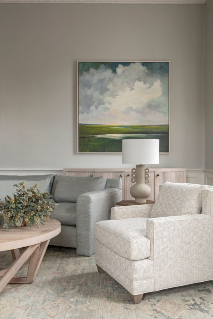 a neutral colored living room designed with GDC Home's in-home interior design consultations in Charleston, featuring an off-white textured armchair, a soft grey sofa, a coffee table and sideboard in the same light wood tone, and a decorative table lamp