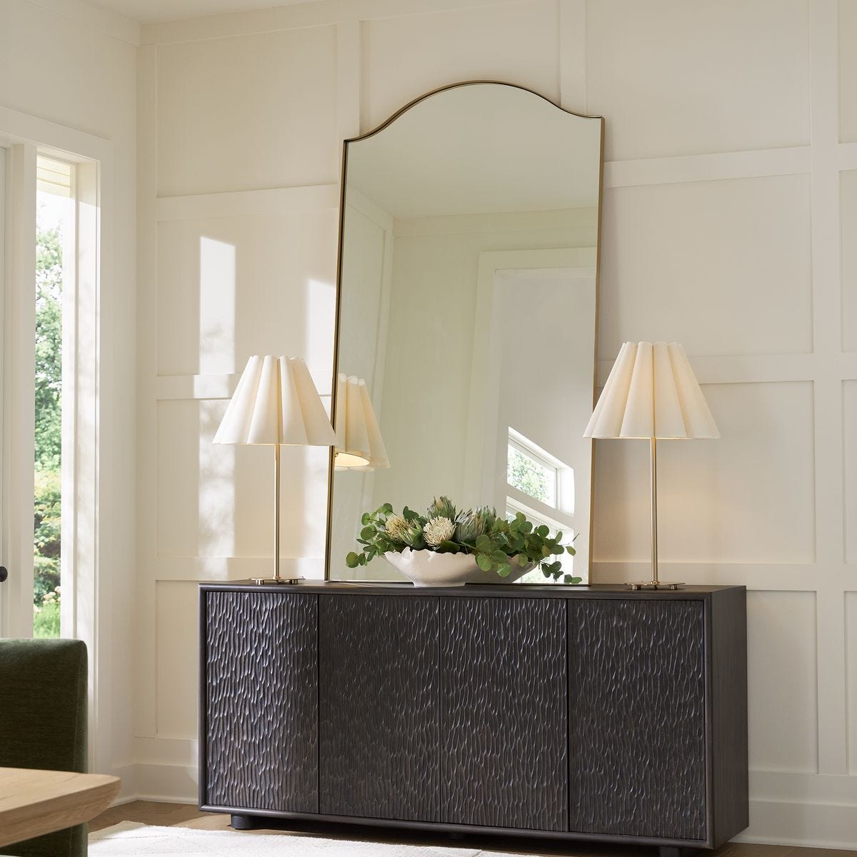 lifestyle image of two brass lamps with white scalloped shades and a large mirror on a console