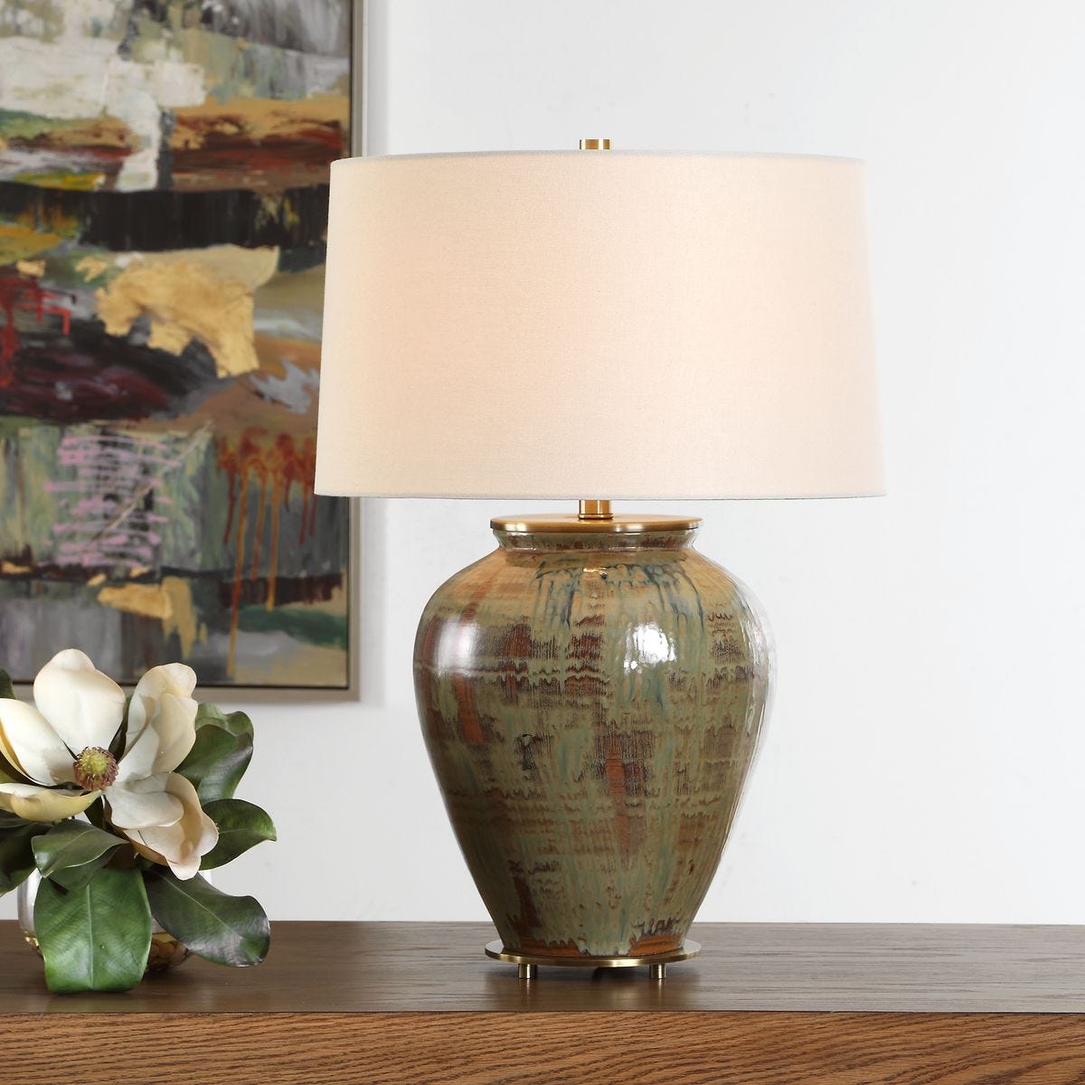 lifestyle image of table lamp with urn base finished in shades of green, blue-green, and earthy browns and an off white shade