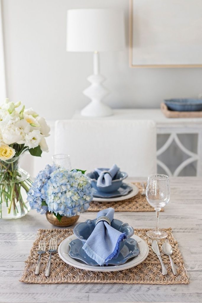 A coastal dining room place setting with delicate woven placemats, silverware and dishware, blue formal napkins, and floral table decor, styled with pieces from GDC's Charleston home decor stores