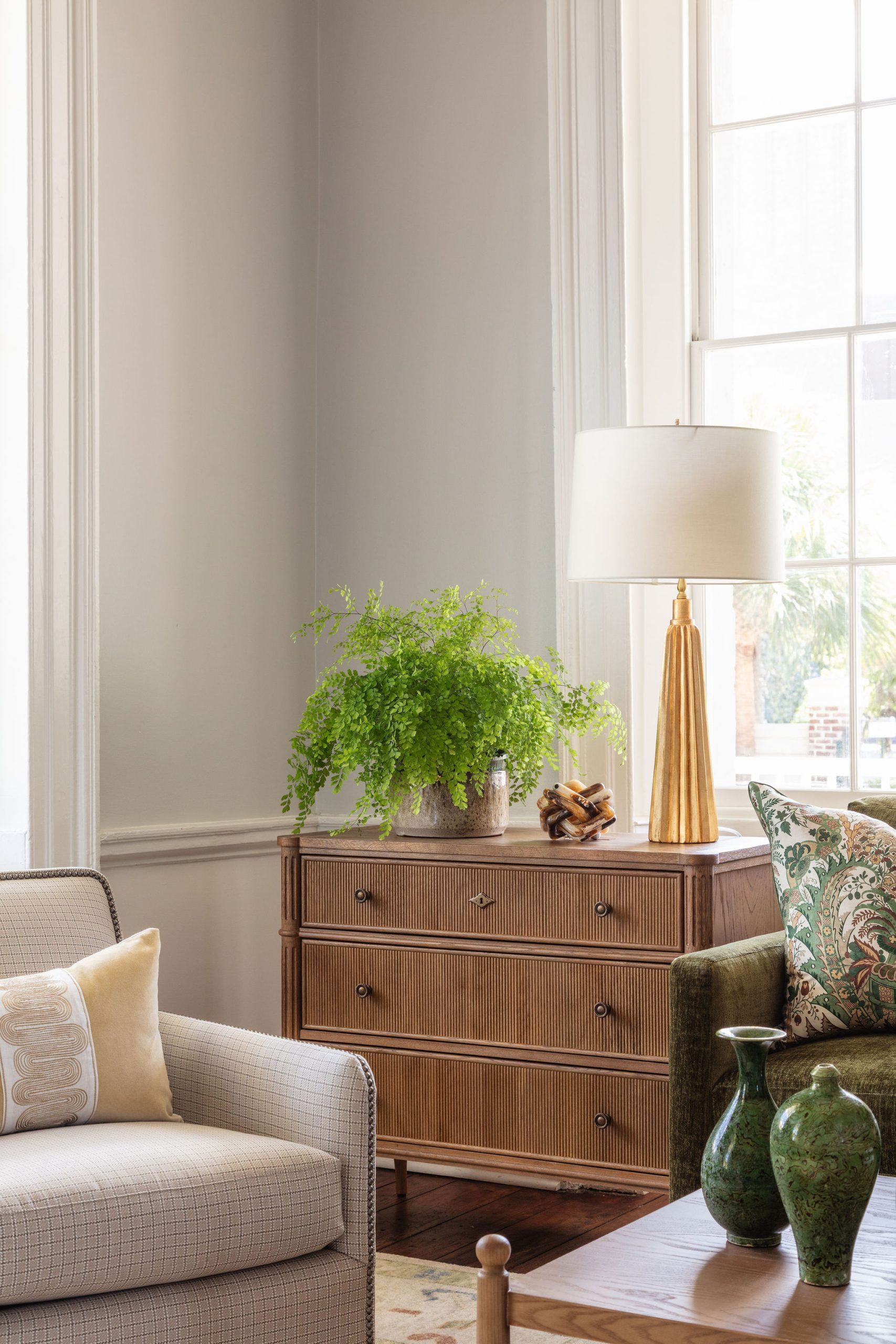 lifestyle image of oak chest with pencil ribbed drawer fronts, gold lamp, greenery, neutral chair, and green vases