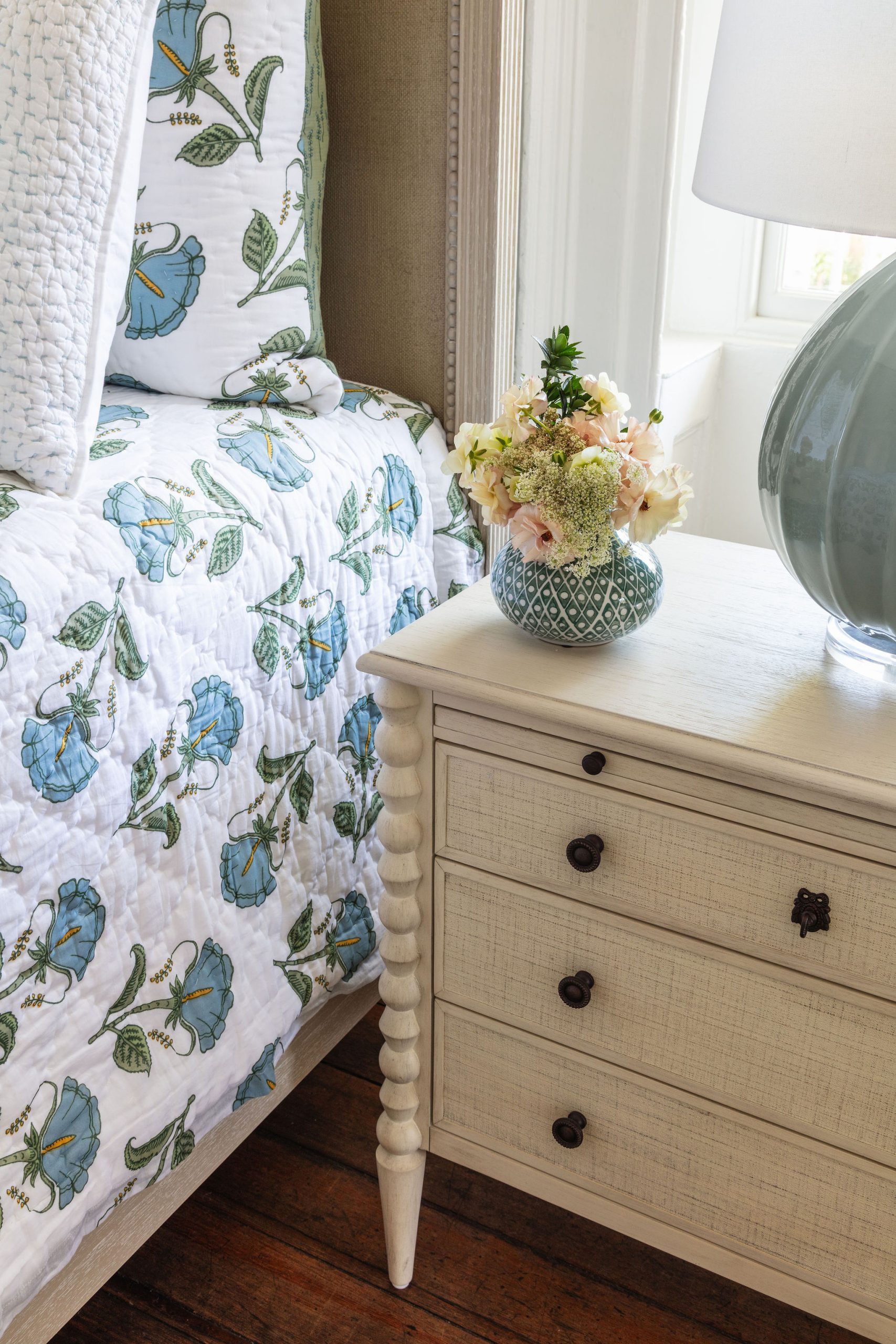 lifestyle image of nightstand with linen front drawers in a chalk white finish, block print bedding, and flowers in a vase