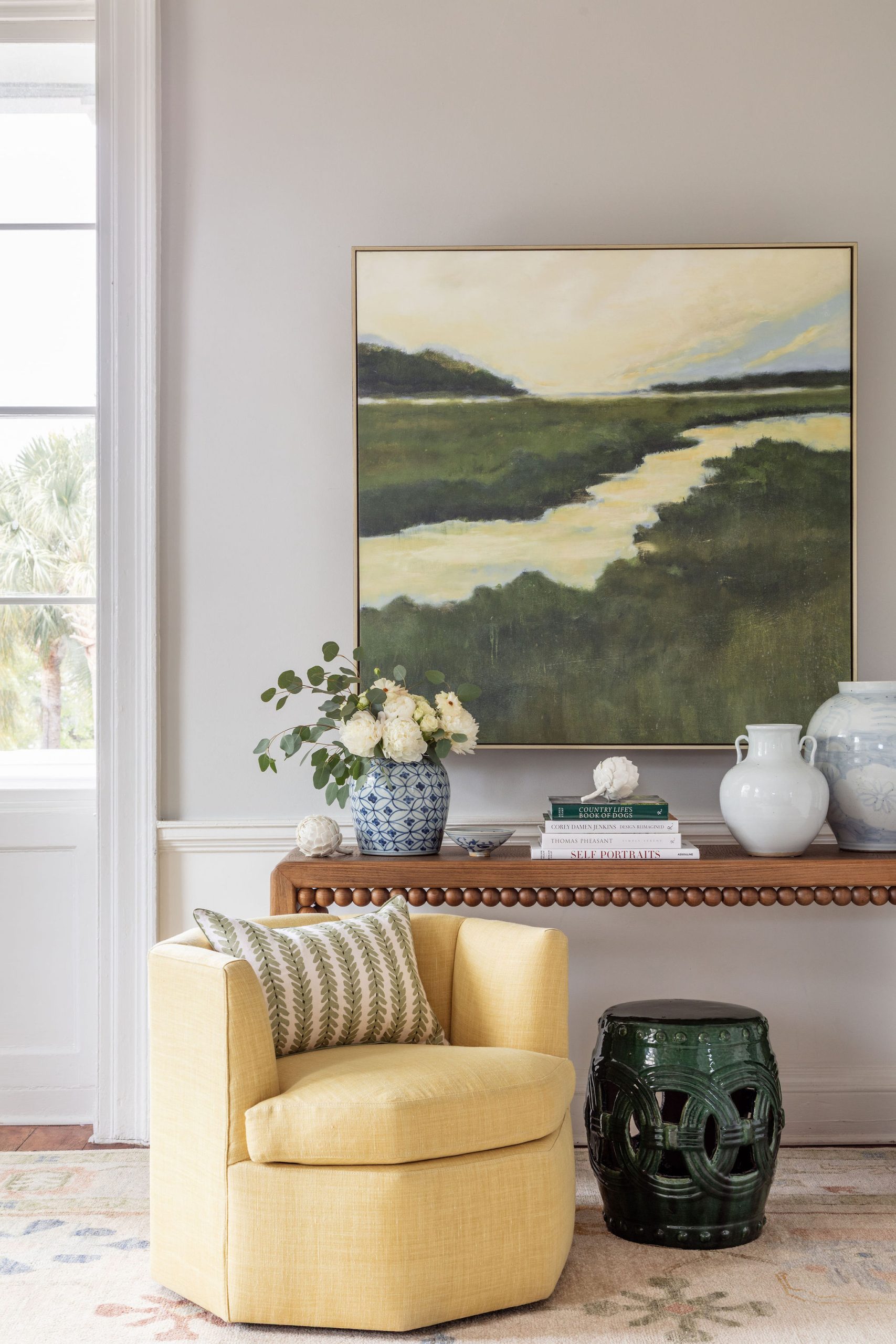lifestyle image of bobbin trim console table with blue and white vases, stack of books, green marsh painting, and a yellow accent chair