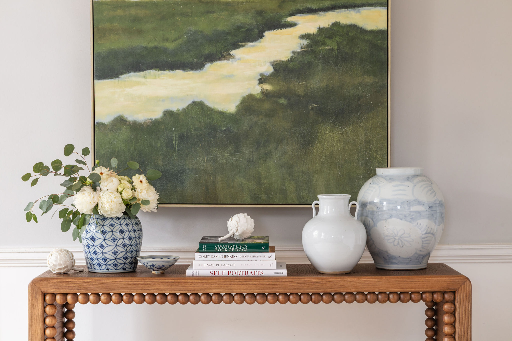 lifestyle image of bobbin trim console table with blue and white vases, stack of books, and a green marsh painting
