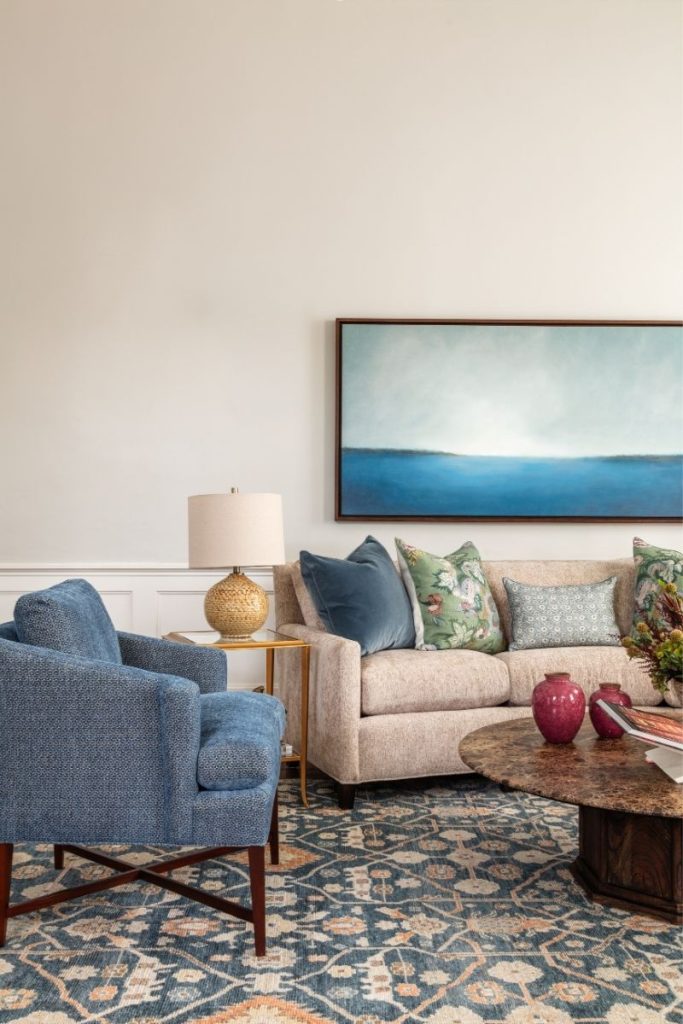 A pop of color accent chair in blue that shares a common color with the sofa's throw pillow, the coastal art, and the rugs in Charleston, SC