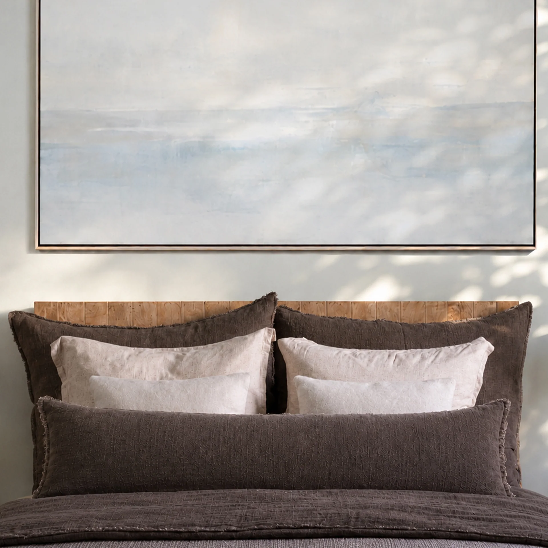 lifestyle image of a bed made up in dark brown linen bedding with blush linen shams and a neutral abstract painting above