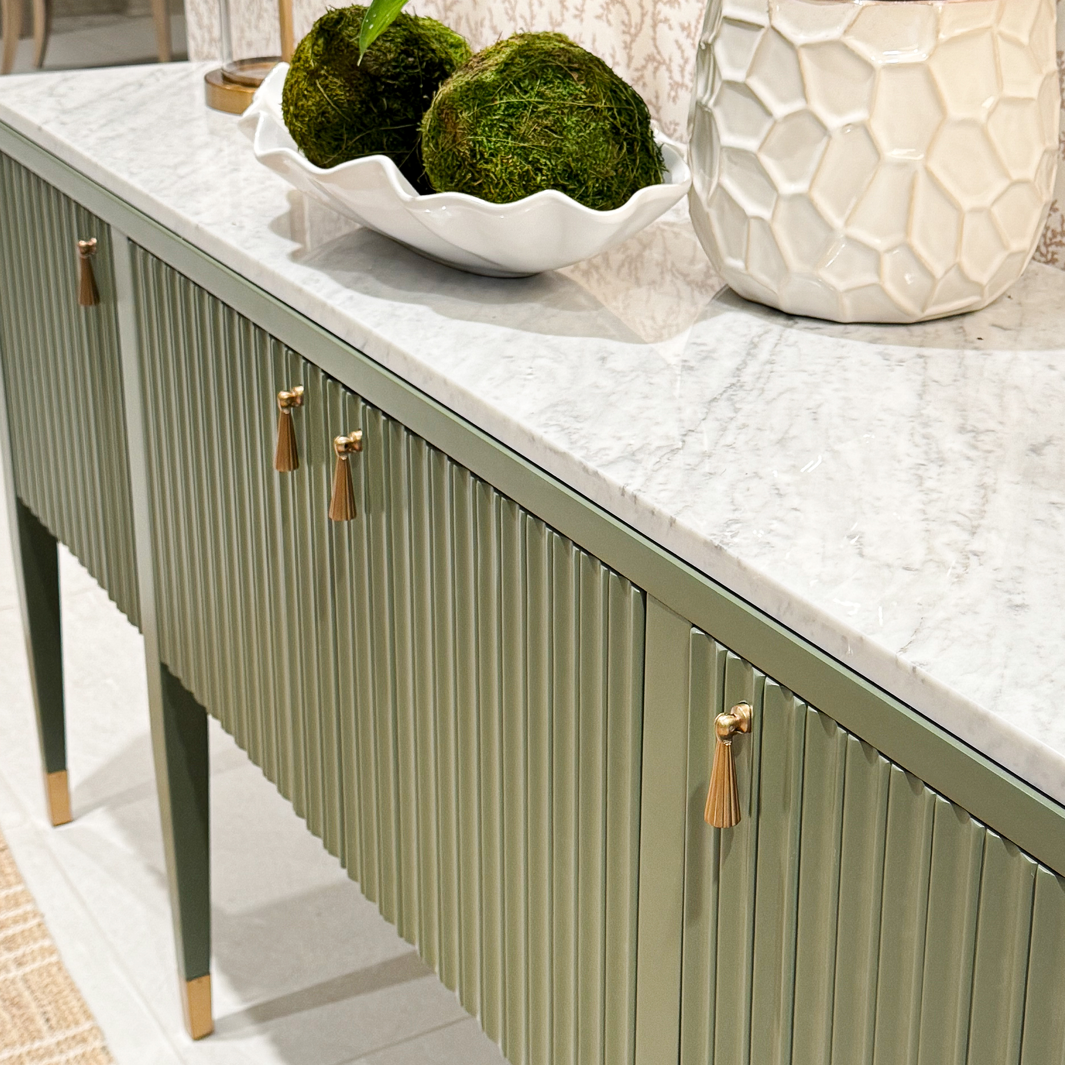 detail of a green console with a marble top, pleated doors, and brass tassel pulls shown with decorative accessories
