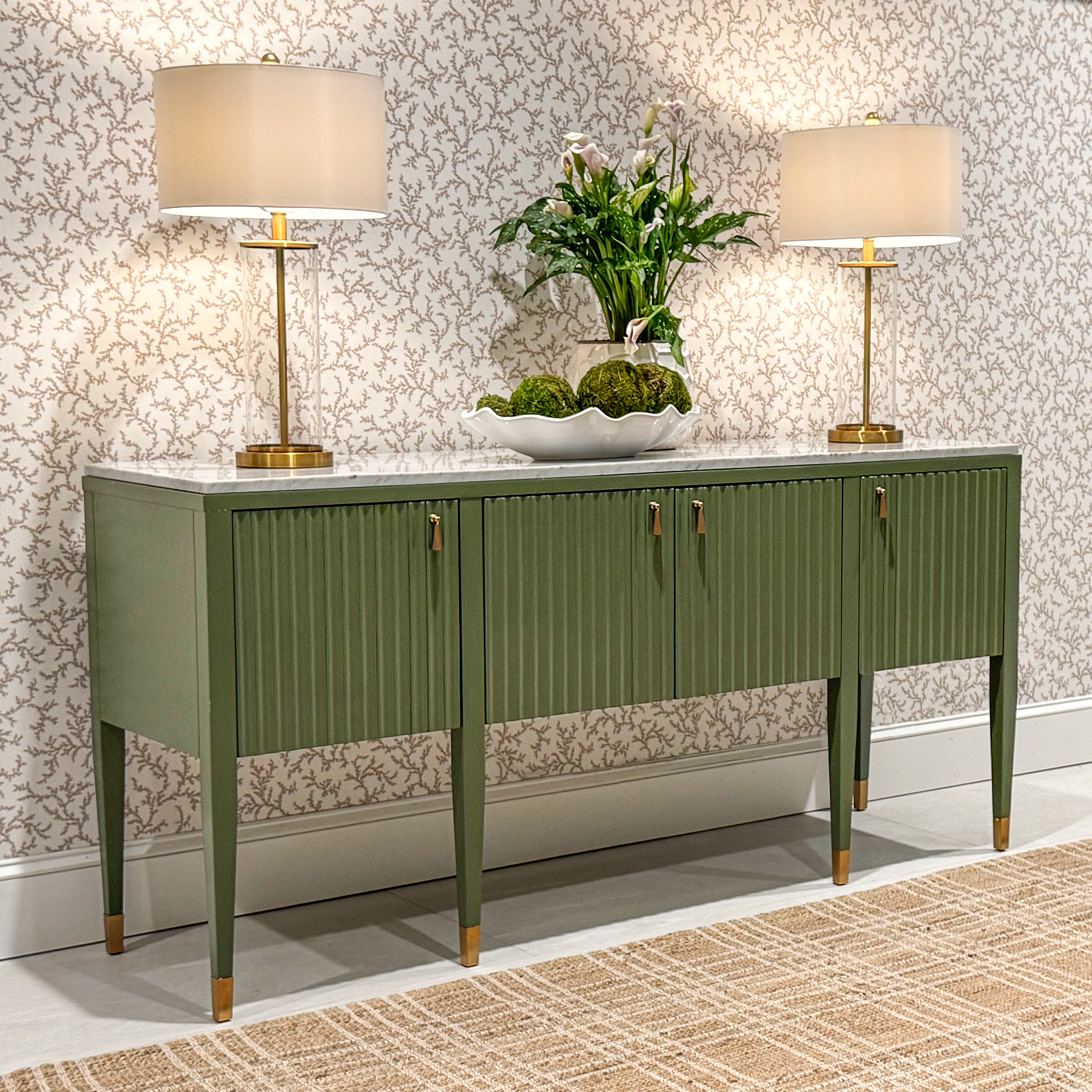 green console with a marble top and pleated doors shown in a wallpapered room with gold buffet lamps and greenery